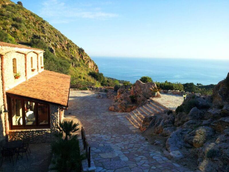 Casale Corcella is a charming stone building and patio in Scopello, overlooking the sea and surrounded by rocky hills and lush vegetation under a clear blue sky.