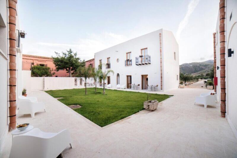 A modern white accommodation with balconies surrounds a courtyard featuring a green lawn, palm trees, white outdoor chairs, and a paved walkway. Mountains rise in the background near Baglio Scopello, Scopello Sicily.