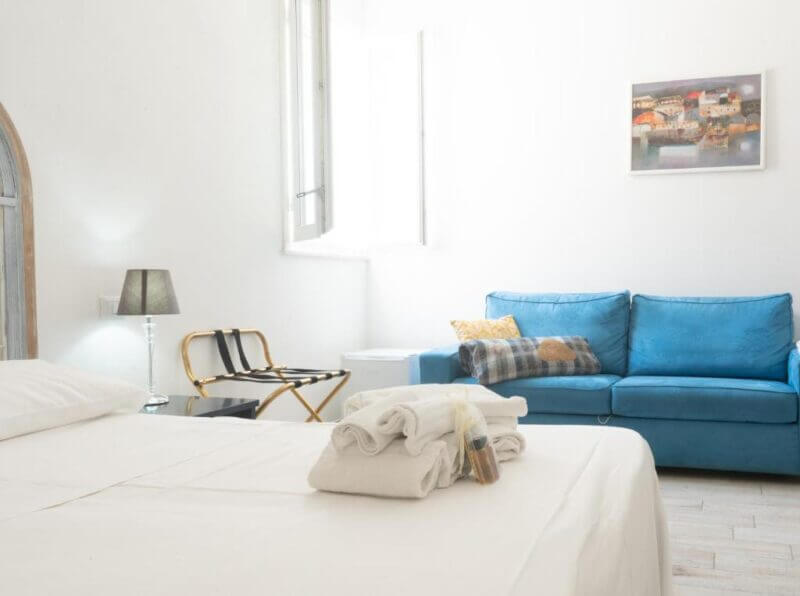 Bright Favignana bedroom with a neatly made bed, folded towels, blue sofa, open window, small lamp, luggage rack, and a framed picture on the wall create a welcoming Poggiamico ambiance.