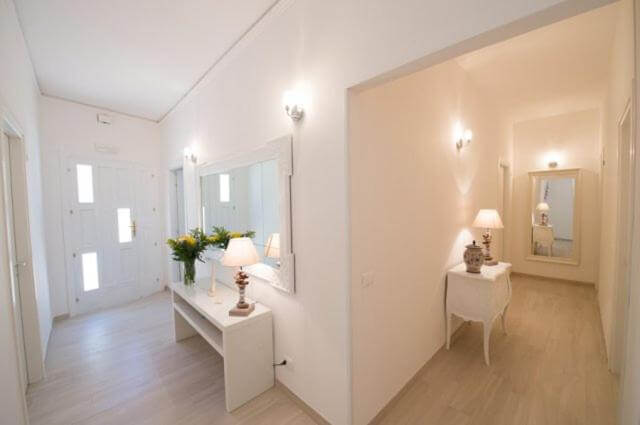 A bright, modern hallway with light-colored walls and floors, decorated with mirrors, lamps, and vases of flowers on white tables, inspired by the serene elegance of Donna Maria Favignana.
