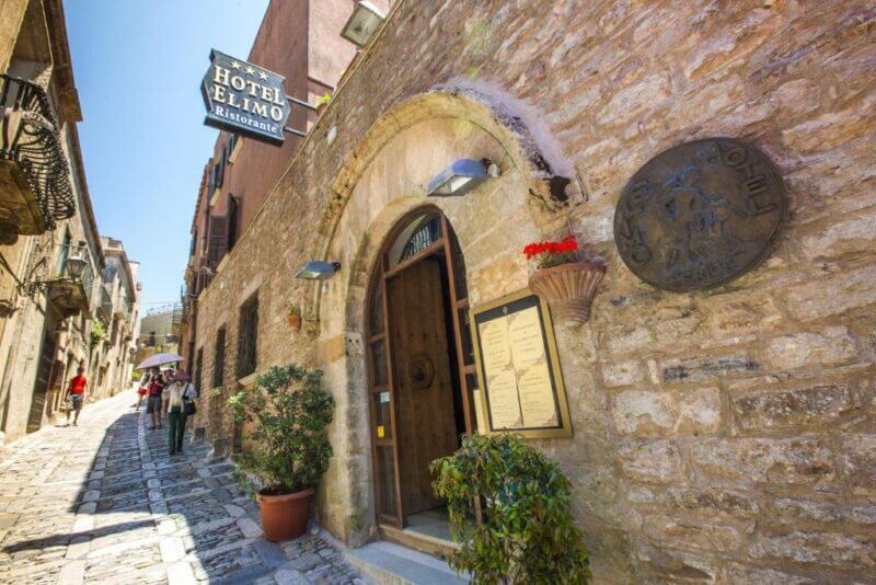 Stone facade of Hotel Elimo, a charming Erice hotel, with arched doorway, menu display, and potted plants on a sloped cobblestone street where people walk uphill.