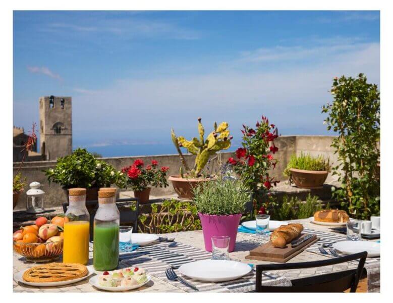 A table set for an outdoor meal at Bella Vista b&b in Erice, with juice, bread, pastries, and fruit; potted plants and a stone building in the background under a clear sky.