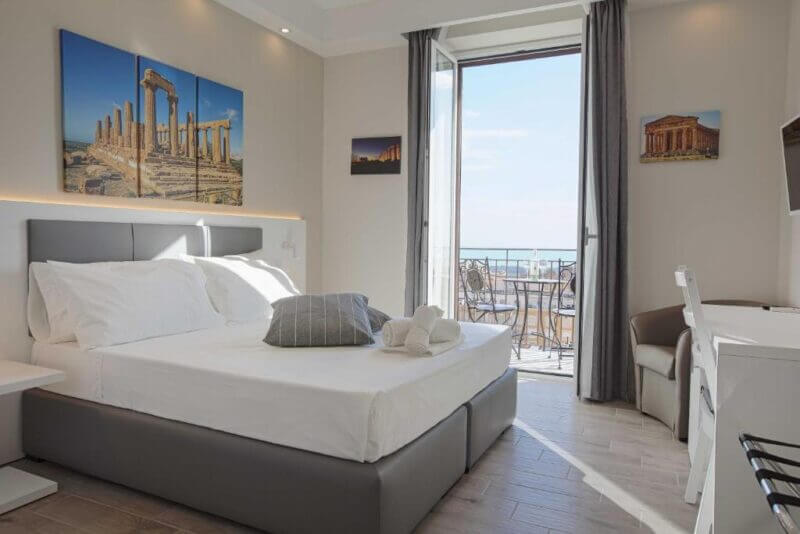 Modern Esseneto hotel room with a double bed, wall art of ancient ruins, desk, and an open balcony door revealing a sea view and outdoor seating in Agrigento.