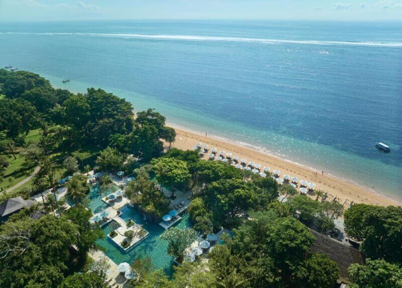 Aerial view of Hyatt Regency Sanur, a Bali resort featuring a large pool, sun loungers, umbrellas, and lush greenery beside the sandy beach and calm blue ocean.
