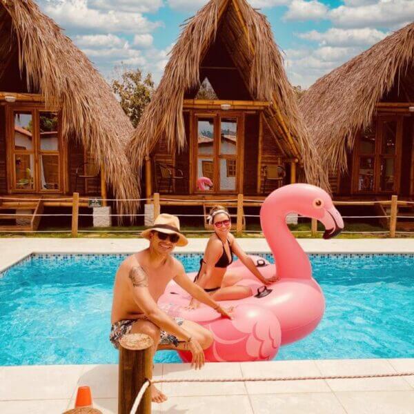 Two people are at a pool with one relaxing on a large pink flamingo float, in front of the charming wooden bungalows of Villa Eden Palomino, their thatched roofs nestled under a partly cloudy sky.
