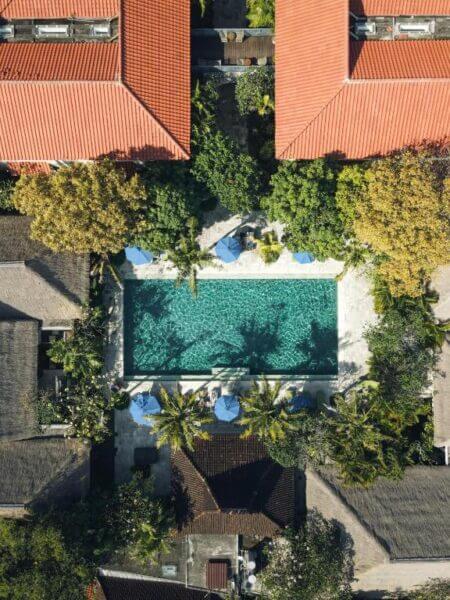 Aerial view of the rectangular swimming pool at Sudamala Resort in Sanur, surrounded by lush trees, lounge chairs with blue umbrellas, and buildings with red and thatched roofs.