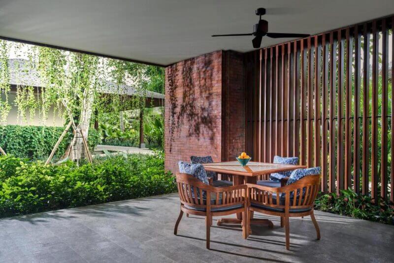 A covered patio at Andaz Hotel in Sanur features a wooden table with four chairs, a ceiling fan above, surrounded by lush greenery, a brick wall, and vertical wooden slats.