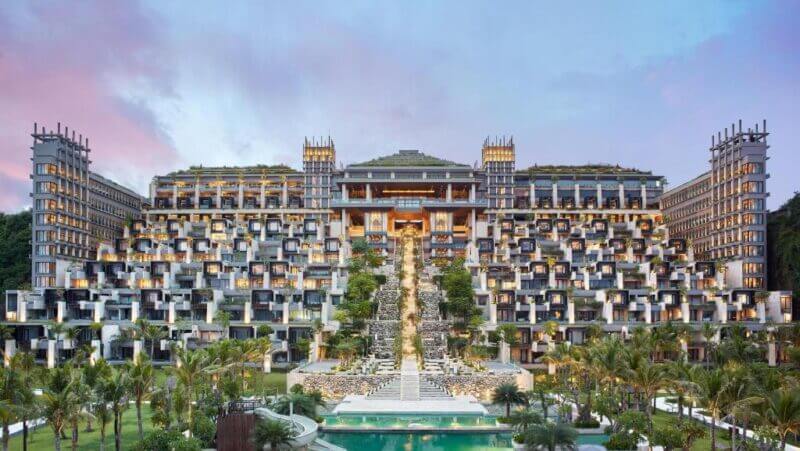 A large, multi-level hotel like Apurva Nusa Dua with terraced balconies features a central grand staircase, surrounded by palm trees and a pool, under a colorful sky.