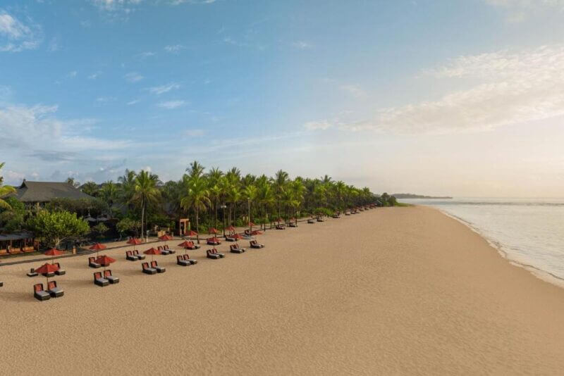 Wide sandy beach lined with red lounge chairs and umbrellas, bordered by palm trees and calm ocean under a partly cloudy sky—pure relaxation at this luxury resort Bali, St. Regis Nusa Dua beachfront hotel.