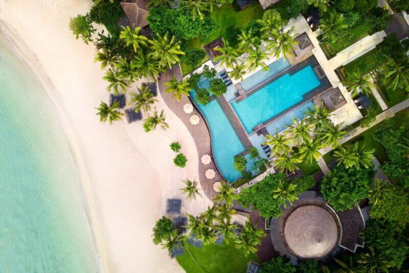 Aerial view of the luxury resort Ritz-Carlton Nusa Dua, featuring a swimming pool, palm trees, lounge chairs, and white sand beside the clear turquoise water.