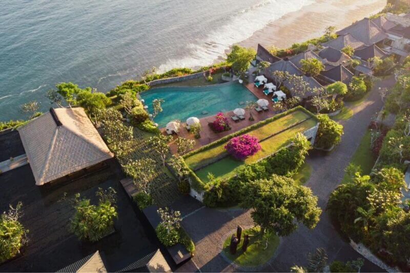Aerial view of Bulgari Resort Uluwatu, featuring a beachfront swimming pool, sun loungers, lush gardens, and elegant buildings nestled close to the ocean.