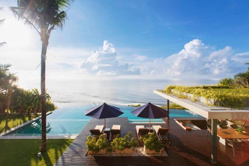 Infinity pool overlooking the ocean with lounge chairs and umbrellas on a clifftop wooden deck in Uluwatu, surrounded by tropical greenery under a clear blue sky.