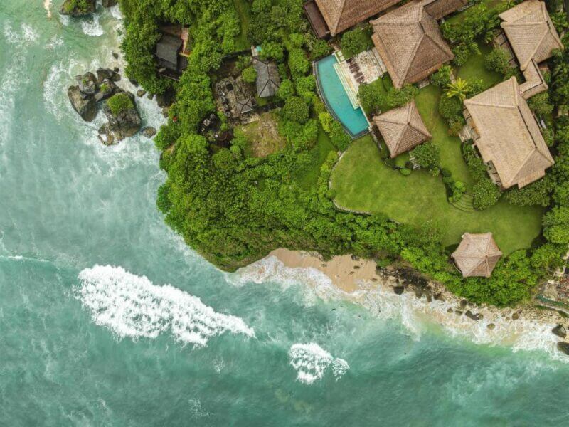 Aerial view of ocean waves hitting a small sandy beach bordered by lush greenery and the luxury resort Radisson Blu Uluwatu, with several elegant thatched-roof buildings nestled along the shoreline.