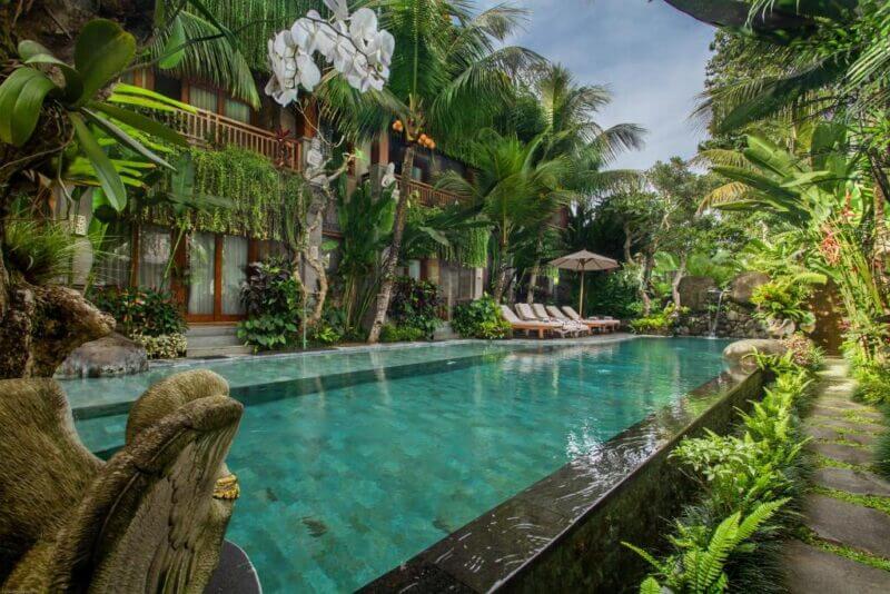 Rectangular outdoor pool at Weda Cita Ubud, surrounded by tropical plants, lounge chairs, an umbrella, and a stone statue, with a multi-story building in the background.