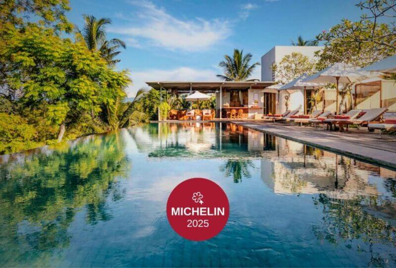 Infinity pool with lounge chairs and umbrellas beside the modern Bisma Eight hotel in Ubud, surrounded by trees under a blue sky. A MICHELIN 2025 badge is overlaid at the bottom center.