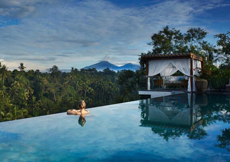 A person relaxes in an infinity pool at Goya Resort Ubud, overlooking a lush forest and distant mountains, with a cabana on the poolside under a partly cloudy sky.
