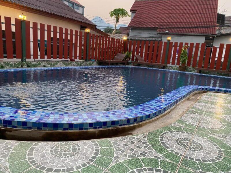 A small outdoor swimming pool with blue mosaic tile edging, surrounded by patterned green tiles and a red wooden fence in the Premier Vang Vieng residential area.