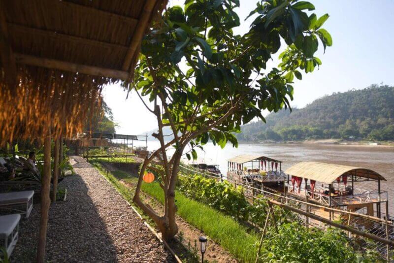 Gravel path beside a shaded seating area, tree, and grassy bank at Belle Rive in Luang Prabang, overlooking the river with docked wooden boats and hills in the background.