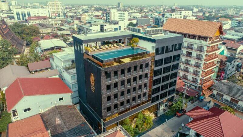 Aerial view of the modern Lao Poet Hotel with a rooftop pool, surrounded by various residential and commercial buildings in urban Vientiane.