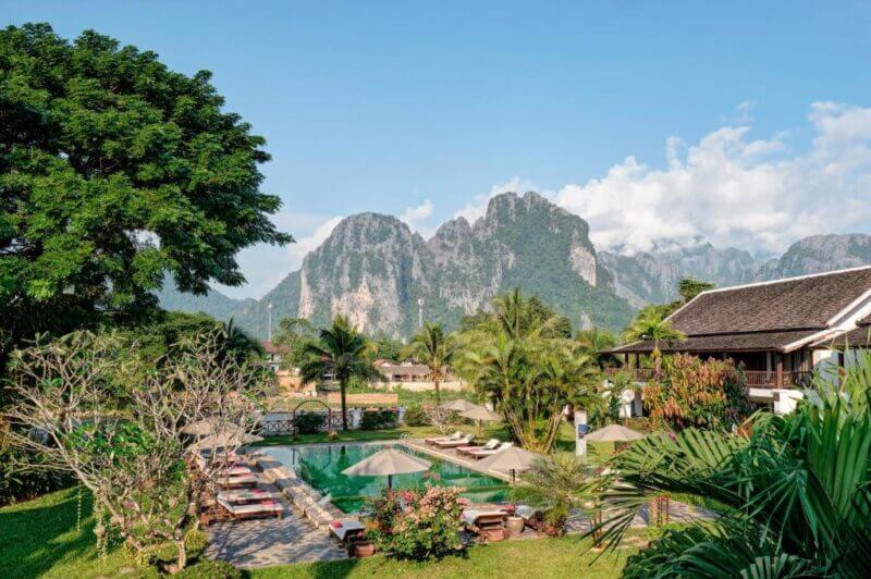 Riverside Boutique Resort in Vang Vieng features a swimming pool, sun loungers, and umbrellas surrounded by greenery, with large mountains rising in the background under a clear blue sky.