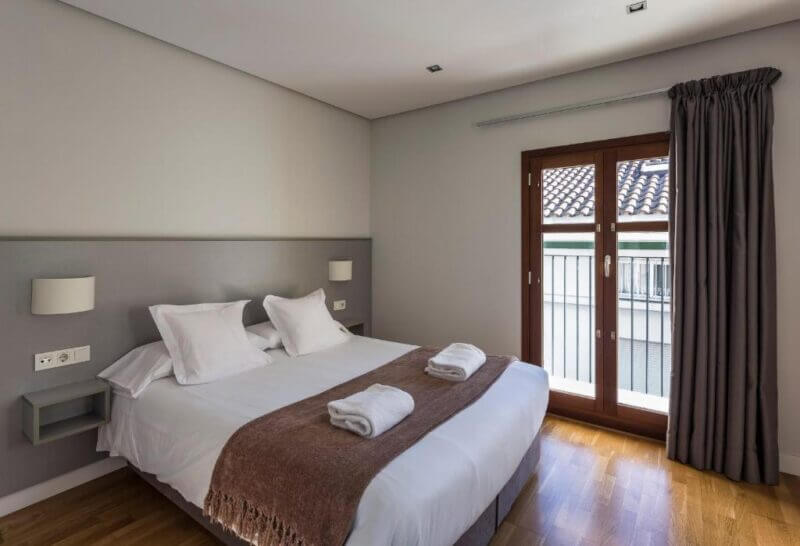 Modern bedroom at Mon Suites Valencia featuring a double bed, two wall-mounted lights, folded towels on the bed, wooden floor, and a window with dark curtains overlooking a tiled roof.