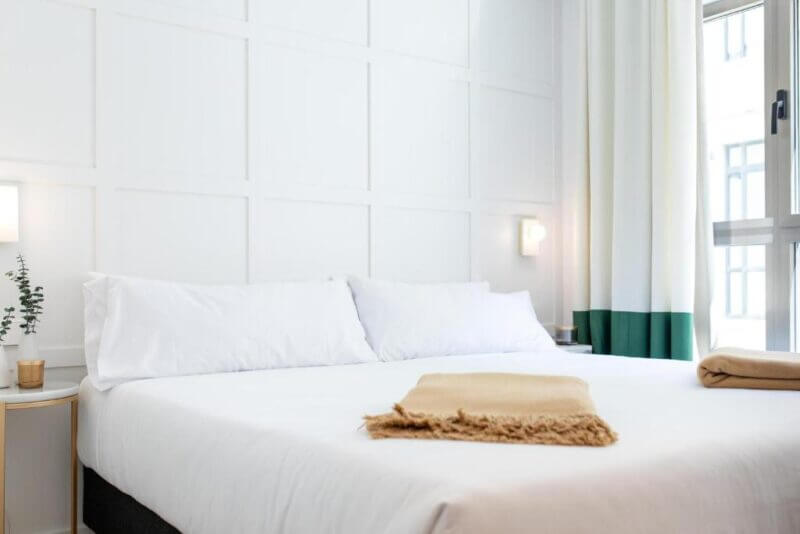 A neatly made bed with white linens, two beige blankets folded at the foot, a nightstand with a small plant, and a window with white curtains in a bright, modern Cosmo Hotel Valencia room.