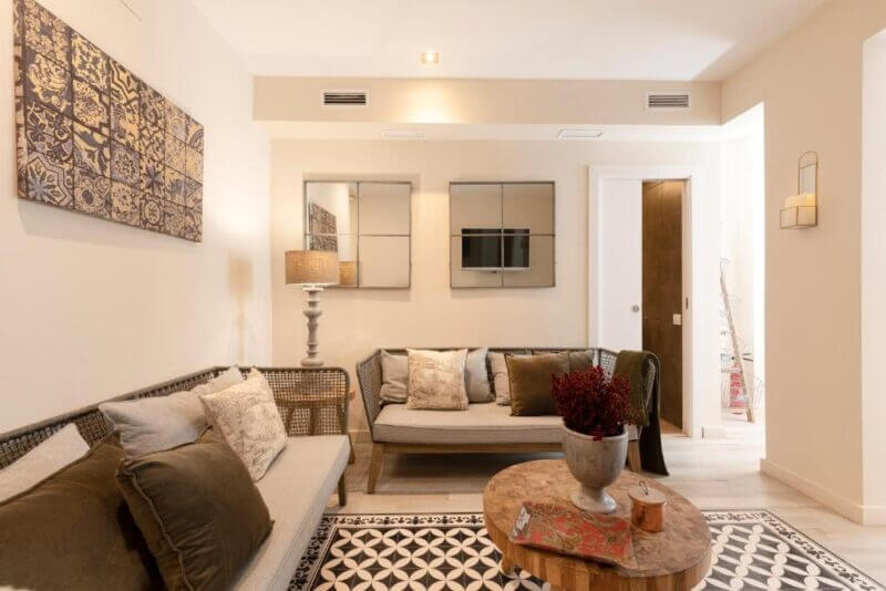 A modern living room in Barcelona with beige walls, a patterned rug, two chic sofas with cushions, a wooden coffee table, decorative wall art, table lamp, and a plant centerpiece—perfect for stylish apartment living.