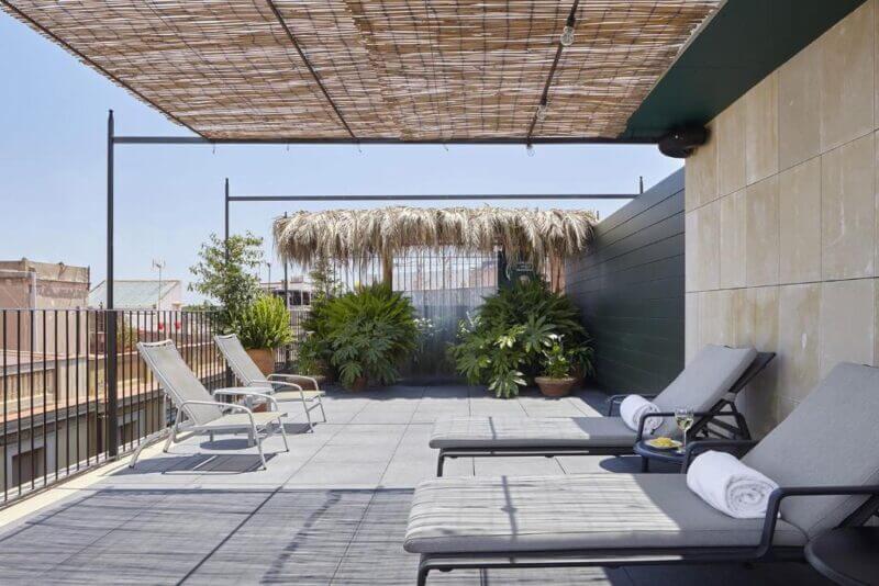 Outdoor terrace at Casa Camper Barcelona with lounge chairs, potted plants, a straw roof shade, and a view of nearby buildings under a clear sky.