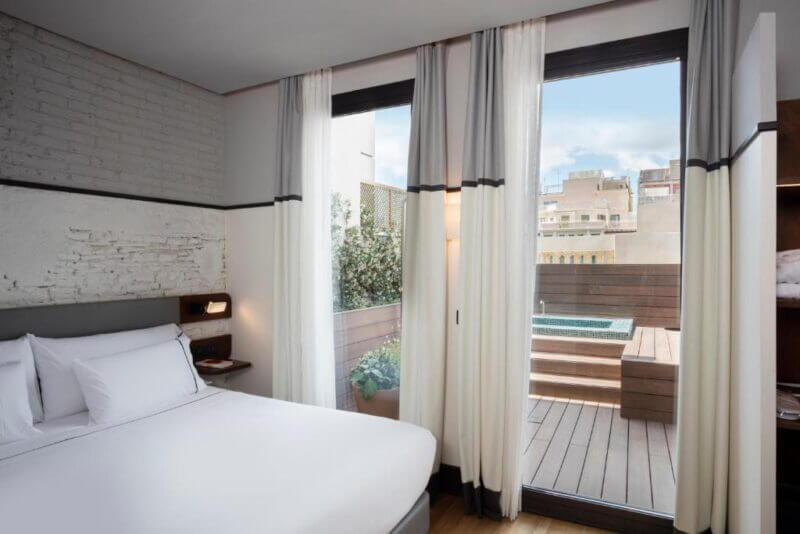 A modern hotel room at Praktik Èssens in Barcelona with a neatly made bed, white curtains, and a private outdoor terrace featuring a small hot tub and city views.