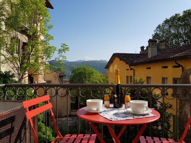 A red table at the Guest House with two cups, juice bottles, and a champagne bottle sits on a balcony overlooking the green hills and yellow buildings of Bergamo Alta under a clear sky.