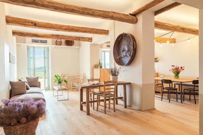 Bright, minimalist living and dining area with wooden beams, light wood flooring, simple furniture, and large windows letting in natural light—perfect for enjoying serene Montepulciano views at CUGUSI.