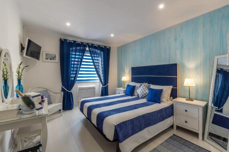 Modern bedroom with blue and white decor, a double bed, nightstands, a vanity, a wall-mounted TV, and a large window with blue curtains inspired by the coastal charm of Ferragudo.