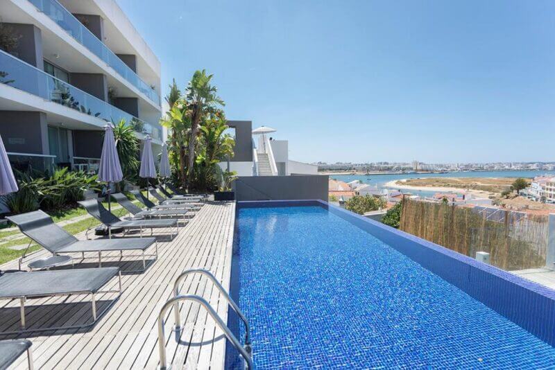 Modern outdoor infinity pool with blue tiles, surrounded by lounge chairs and umbrellas, next to One2Seven Ferragudo—a multi-story hotel with balconies—overlooking a coastal town and bay for the perfect Algarve holiday.