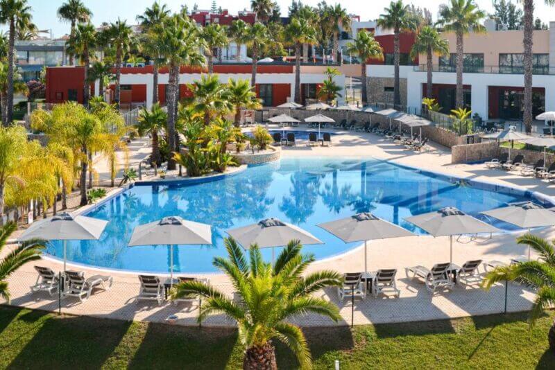 Spacious outdoor swimming pool at Vitor's Village, surrounded by palm trees, lounge chairs, and umbrellas, with the modern hotel building in Ferragudo providing a stunning backdrop.
