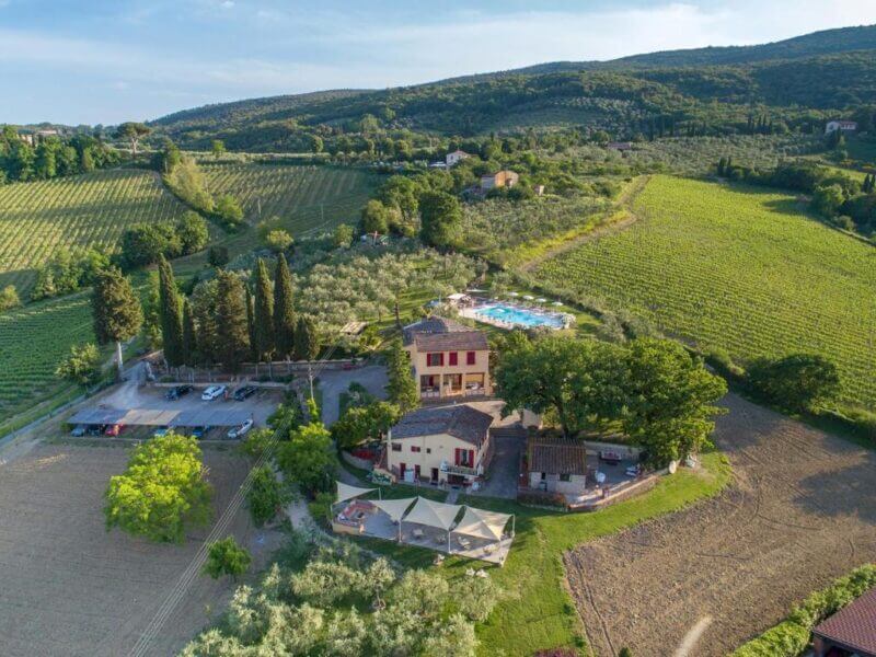 Aerial view of Bellavista, a countryside estate near San Gimignano, featuring several buildings, a swimming pool, parking area, and surrounding vineyards and trees under a clear sky.
