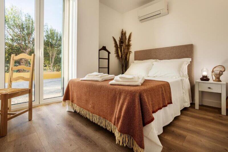 A neatly made double bed with a brown blanket and white pillows sits in a bright Agro Turismo room with wooden floors, a nightstand, chair, and large glass doors opening to the outside, offering an inviting stay in Evora.