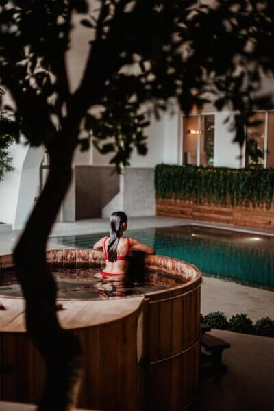 A woman in a red swimsuit sits on the edge of a round wooden hot tub at Lamego Hotel, near an indoor pool, with greenery and modern decor evoking the serene landscape of the Douro in the background.