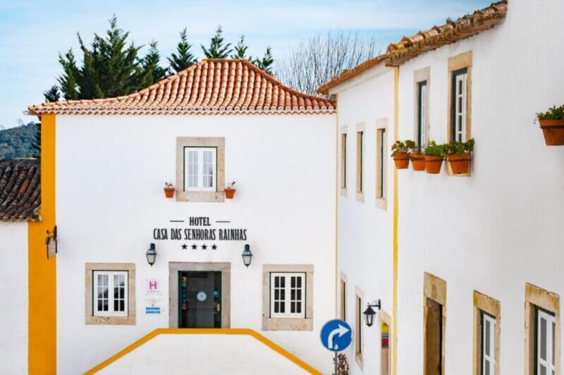 White hotel building with orange roof tiles labeled Casa Senhoras Rainhas in Obidos, flanked by windows with flower boxes and a blue one-way traffic sign in front.