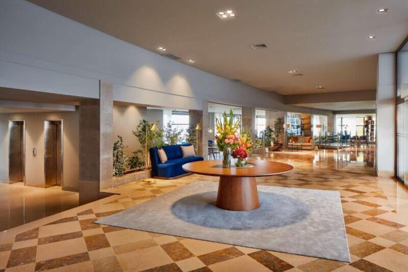 Spacious hotel lobby with a round wooden table topped with floral arrangements, marble flooring, elevators on the left, and seating areas in the background.