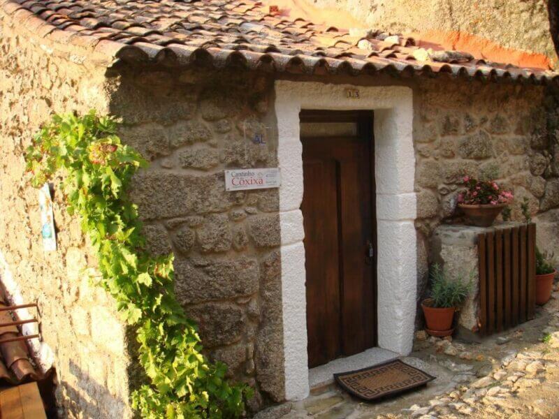 A small stone house with a wooden door, white trim, potted plants, and a sign reading Cozinha next to the entrance—this charming spot in Monsanto is known as Cantinho da Coxixa.