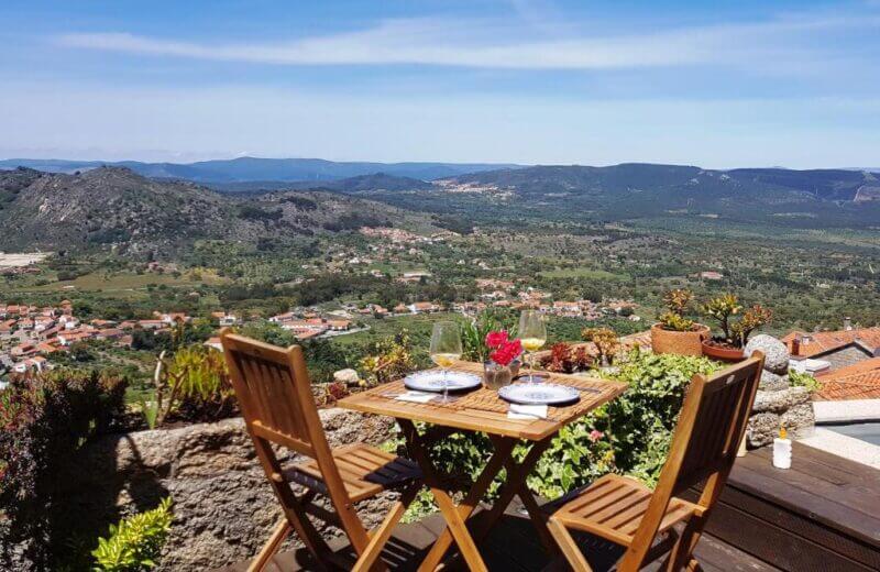 A wooden table with two chairs set for a meal overlooks a scenic valley in Monsanto, with mountains, a village, and Casa do Castello Monsanto visible under a clear blue sky.