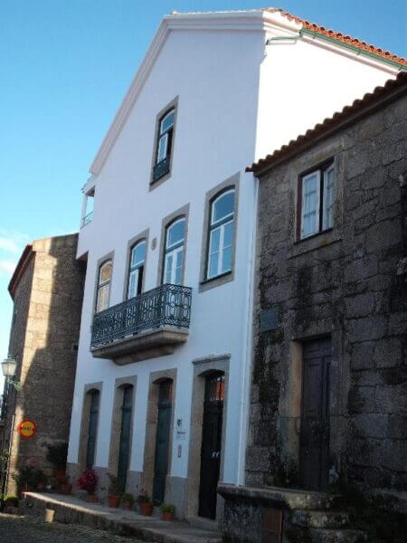 A white, two-story building—Casa Pires Mateus—with a balcony stands beside an older stone building on a cobblestone street in the historic village of Monsanto, under a clear blue sky.