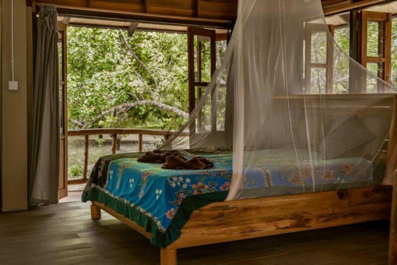 A wooden bed with a colorful floral bedspread and mosquito net sits in a room with open windows and doors at Monkey Mansion Khao Sok, overlooking green trees outside.
