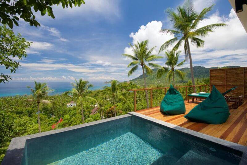 Infinity-edge pool with two green bean bag chairs on a wooden deck overlooks lush tropical trees, hills, and the ocean under a blue sky at Overthemoon Koh Tao—a stunning Koh Tao hotel retreat.