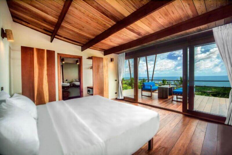A modern bedroom at Ecotao Lodge on Koh Tao features wood floors and ceiling, a large bed, and sliding glass doors opening to a balcony with stunning ocean and sky views.