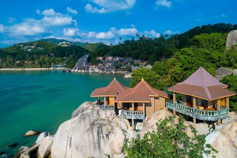 Overwater villas with red roofs are built on rocks along a tropical coastline at Koh Tao Freedom Beach, with turquoise water, lush green hills, and scattered clouds in the sky.