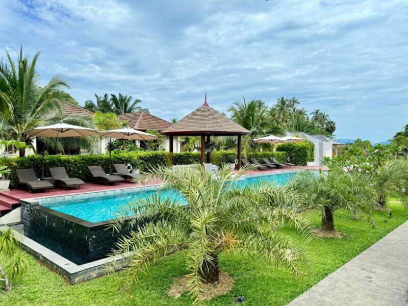 A rectangular outdoor swimming pool with lounge chairs, umbrellas, and palm trees awaits at Verona Koh Lanta, set in a tropical garden under a partly cloudy sky.