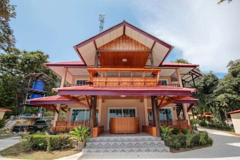 A two-story hotel with a red roof, wooden accents, large front porch, and steps leading to the entrance, surrounded by plants and trees—perfect for a relaxing stay at OYO 1085 in Koh Lanta.