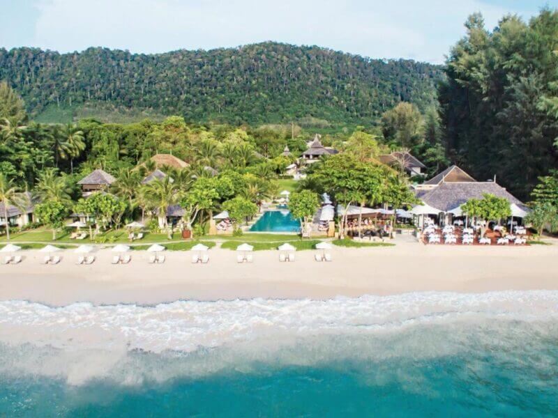 A beachfront resort on Koh Lanta with white sand, lounge chairs, a pool, and tropical greenery sits at the edge of a forested hillside.