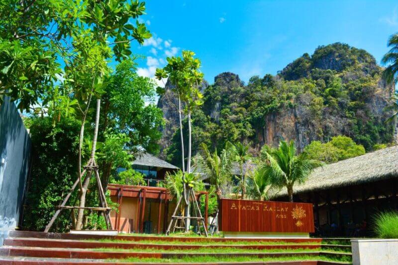 A landscaped area with young trees, a thatched-roof building, and a wooden sign reading AYARA KAMALAY at Avatar Railay, Krabi, set against rocky, forested hills and a brilliant blue sky.